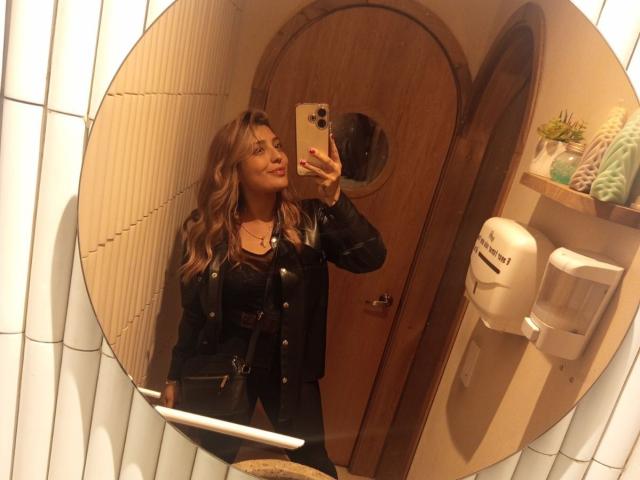 BiancaRey with wavy blonde hair, black leather jacket, and necklace, taking a selfie in a mirror, showing off her tits, in a warmly lit, tiled bathroom.