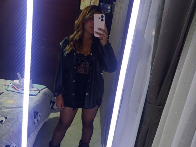 BiancaRey stands in front of a mirror, taking a selfie with an iPhone. She wears a black leather jacket, sparkly top, and black shorts, with black ankle boots.