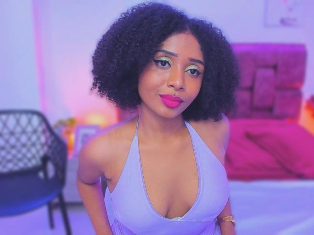 MeganWilliams with curly black hair, wearing a purple halter top, showing her boobs, pink lipstick, green eye makeup, and a gold watch, smiling in a brightly lit room.