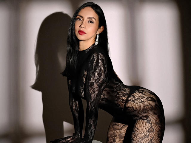 BelladonnaLips, en body noir en dentelle, dévoilant ses seins et ses fesses, à genoux, regardant la caméra, lèvres rouges, cheveux noirs, ombre sur le mur, boucles d'oreilles, sexy, confiante..