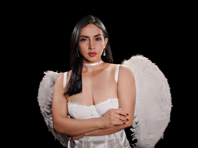 BelladonnaLips avec des ailes d'ange blanches, des cheveux noirs, un corset en dentelle blanche, les bras croisés, des ongles rouges, un collier ras du cou et une expression sérieuse, fond noir. Les seins, les mamelons et les parties génitales ne sont pas visibles..