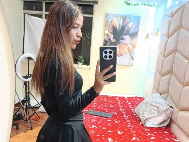 ValerieWoods met lang bruin haar, in een zwarte jurk, die een spiegelzelfportret maakt, waarbij haar achterkant te zien is, terwijl ze een zwarte iPhone vasthoudt, in een kamer met een rode sprei op het bed..