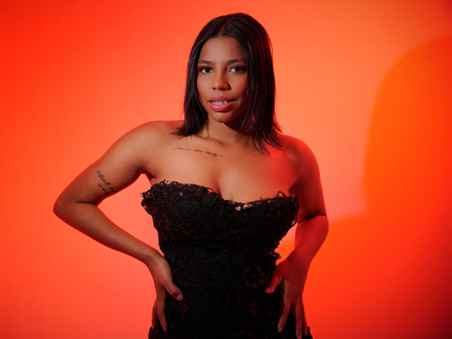 SammyRosi with black lace dress, hands on hips, medium brown skin, black hair, tattoos on arm, pink lipstick, red-orange background, and confident expression. Boobs visible, tits pushed up.