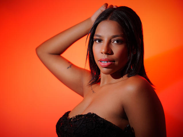 SammyRosi with dark skin, black lace strapless top, blue lipstick, black hair, arm raised, tattoo on bicep, looking at camera, red background, confident, sexy, attractive.