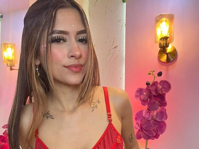 AfroditaWild with medium brown hair, red lipstick, and a nose piercing, wearing a red lace bra, has tattoos on her chest and eyeliner. Bright wall scones and pink orchids.