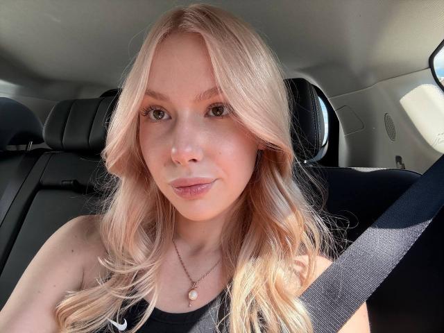 Laurinda Lupacchino hat blondes Haar, trägt ein schwarzes Oberteil, einen Sicherheitsgurt und eine Halskette mit einem rosafarbenen Schmuckstein. Sie sitzt in einem Auto, lächelt und trägt dezentes Make-up mit einem natürlichen Hautton..