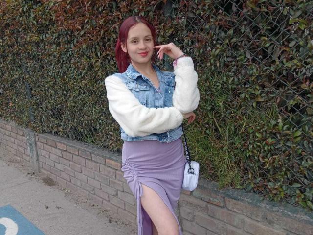 LesleyGray with red hair, denim vest, white fuzzy sleeves, purple skirt with thigh slit, holding phone, standing by brick wall, white crossbody bag, smiling, cute.