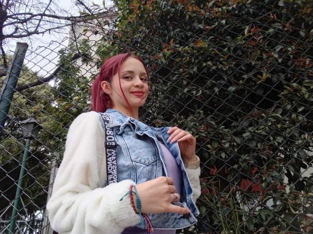LesleyGray with pink hair, white fuzzy sweater, and blue denim vest, standing by a chain-link fence, smiling, showing a small purple bra, holding a colorful bracelet.