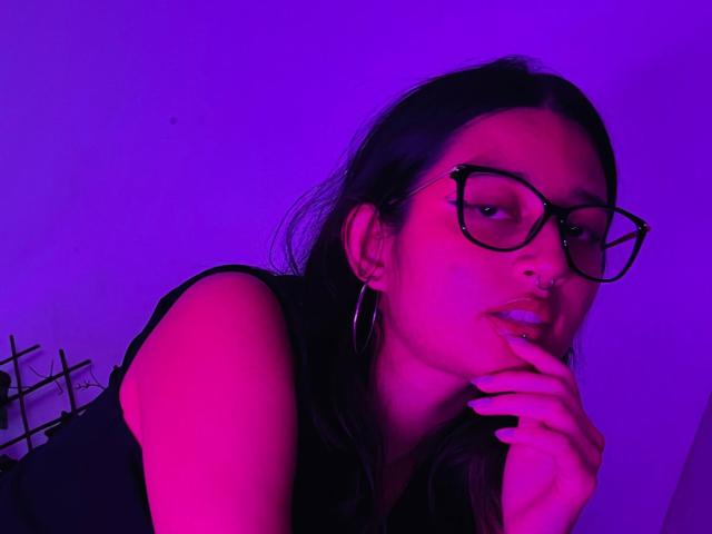 AriaLilith con iluminación rosa-morada, cabello oscuro, piercing en la nariz, gafas grandes, aros, y un dedo tocando sus labios, mostrando ligeramente sus pechos, vistiendo una camiseta negra sin mangas..