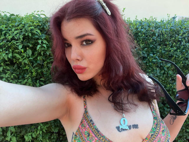 AliceMarina met rood haar, volle lippen, groene ogen, die een kleurrijke beha draagt, zwarte hoge hakken vasthoudt en haar borsten laat zien met een tatoeage van "1 omhoog"..