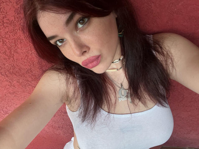 AliceMarina mit roten Haaren, grünen Augen, rosafarbenen Lippen, einem weißen Tanktop, einer Halskette und einem Choker, die ihre Brüste zeigt, vor einer roten, strukturierten Wand..