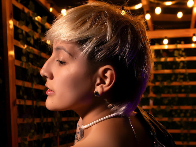 DannyRouds ha i capelli biondi e viola, corti e disordinati, tappi per le orecchie neri e una collana di perle, con un tatuaggio sul collo, sullo sfondo di un ambiente caldo, in legno e con una struttura a griglia..