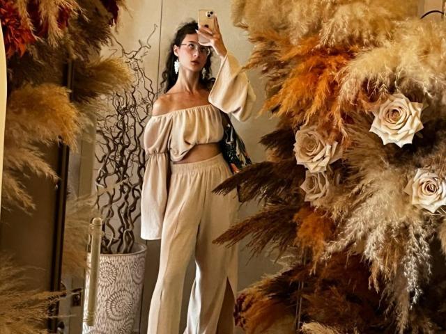 AlyceErway, con i capelli scuri e ricci, gli occhiali, una maglietta bianca a spalle scoperte e pantaloni bianchi a vita alta, sta scattando un selfie allo specchio. È circondata da soffici cuscini marroni e beige e da rose..