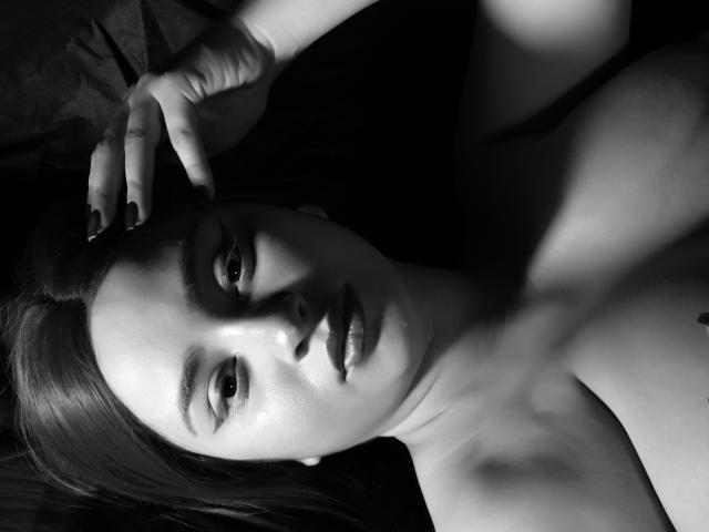 GraceBlue69 ha le spalle scoperte, una mano tra i capelli scuri, unghie nere e uno sguardo serio, con il viso lucido, in bianco e nero, ad alto contrasto, sexy, senza top, attraente e pensieroso..
