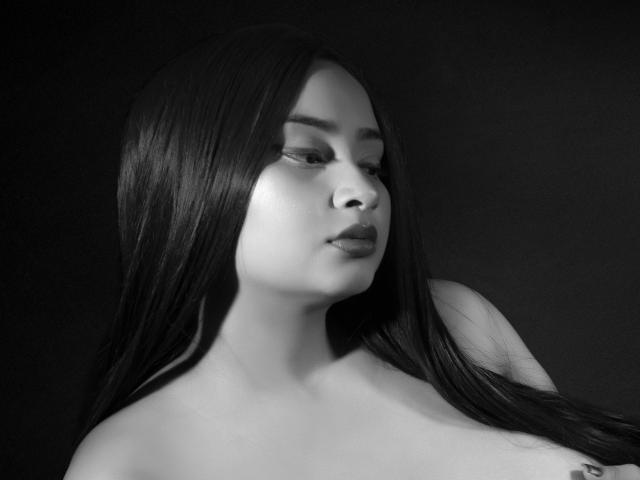 GraceBlue69 met lang donker haar, een serieuze uitdrukking en gladde huid, die lichtelijk opzij kijkt op een zwart-wit foto, waarbij haar blote schouders en een subtiele tatoeage op haar borst te zien zijn..