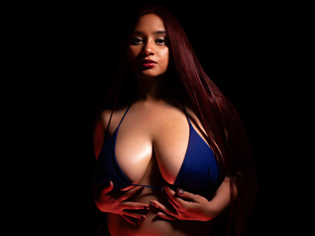 GraceBlue69 con lunghi capelli rossi, bikini blu, grandi seni, mani che coprono i capezzoli, sfondo scuro, illuminazione rossa, espressione seducente, pelle lucida, dettagliata, realistica..