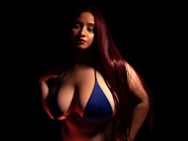 GraceBlue69, avec de longs cheveux roux, un haut de maillot de bain bleu, de grandes seins et un sourire séduisant, sur un fond sombre, avec un éclairage rouge doux qui met en valeur ses courbes..