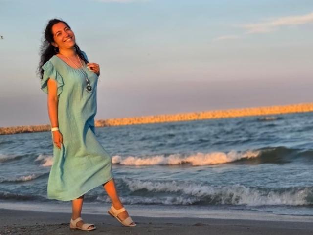 SexyMirra, com cabelos escuros e cacheados, vestindo um vestido verde solto, sandálias brancas e um colar, sorri na praia, estando de pé na areia com ondas suaves ao fundo..