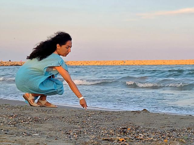 SexyMirra est accroupie sur une plage rocheuse, vêtue d'une robe bleue et de sandales, et touche l'eau avec sa main droite. Elle a les cheveux noirs et bouclés, une peau claire, et la lumière du soir est douce..