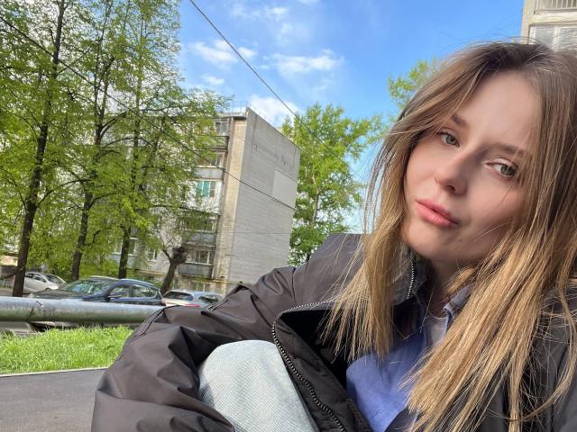 Lera Kreiger mit hellbraunen Haaren, blauen Augen und rosafarbenen Lippen, trägt eine schwarze Jacke und ein blaues Hemd und steht im Freien, blickt in die Kamera, in einer sonnigen, grünen, städtischen Umgebung..