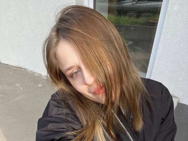 Lera Kreiger, avec des cheveux bruns de longueur moyenne, légèrement en désordre, recouvrant un œil, portant une veste noire, a une peau claire et des yeux verts clairs, et se tient à l'extérieur, souriant légèrement..