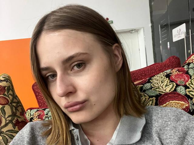 LeraKreiger a une peau pâle, des cheveux bruns clairs et lisses, des yeux verts et une petite tache de naissance sur sa joue droite. Elle porte une chemise grise à col avec des coussins floraux colorés..