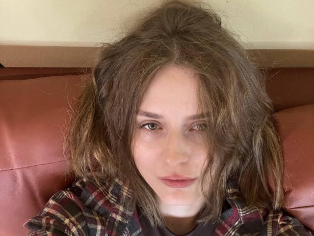 Lera Kreiger, avec ses cheveux bruns en bataille, ses yeux verts et son rouge à lèvres rouge, portant une chemise à carreaux, assise sur un siège en cuir rouge. Son visage est légèrement incliné, avec une expression neutre..