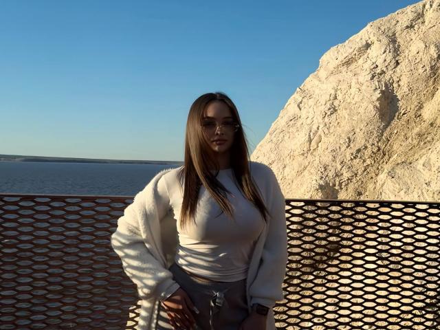 Tawnya Kaczmarski, avec de longs cheveux bruns, portant des lunettes de soleil, un haut blanc et un pantalon gris, se tient devant une clôture en métal, près d'une falaise rocheuse, sous un ciel bleu clair..