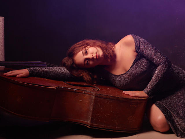 Milana Miller, met golvend bruin haar, draagt een glinsterende zwarte jurk en leunt tegen een houten cello. Ze laat haar borsten zien en heeft een sensuele glimlach, alles in gedimd paars licht..