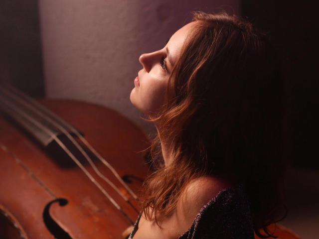 Milana Miller mit welligem, braunem Haar, voller Lippen und einem Profil, die zu einem hölzernen Cello aufschaut, wobei ihre Brüste leicht sichtbar sind, in einem schwach beleuchteten, warmen Bild..