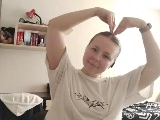 Johnetta Urtiaga, met kort bruin haar, lachend, draagt een wit T-shirt met een zwart boommotief, haar armen omhoog, en maakt met haar vingers een hartvorm, in een helder verlichte kamer..