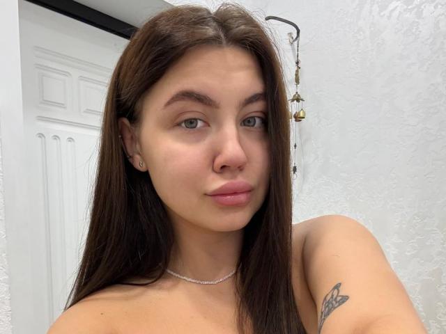 InesGueningsman has long brown hair, green eyes, pink lips, a small nose, a necklace, and a butterfly tattoo on her right arm. She's wearing a sleeveless top.