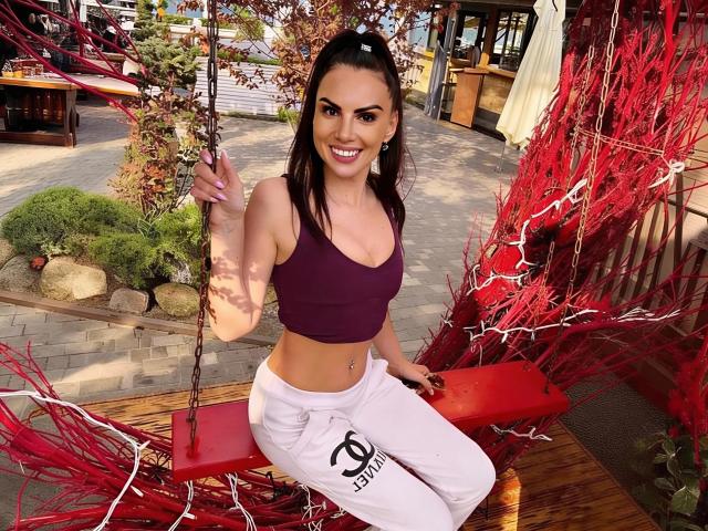 SaraSwede with dark brown hair, smiling, wearing a purple sports bra and white pants, sitting on a red swing with red branches, showing her toned, tan, and cute ass.