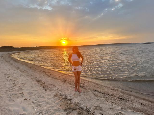 SaraSwede está em uma praia de areia ao pôr do sol, vestindo uma camiseta cropped branca e shorts, com as costas voltadas para a câmera, e sua nádega ligeiramente visível, com o sol atrás dela..