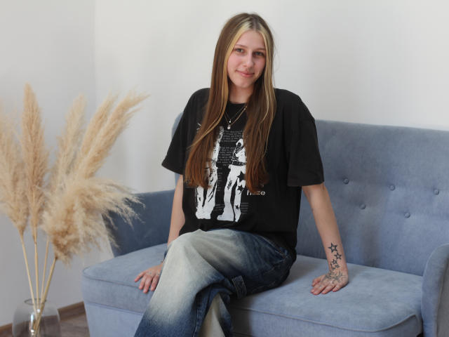 MiaNeit mit langen blonden Haaren, schwarzem T-Shirt, blauen Jeans, sitzend auf einem grauen Sofa, Tätowierung am rechten Unterarm, buschige, braune Gräser auf der linken Seite, lächelt sanft..