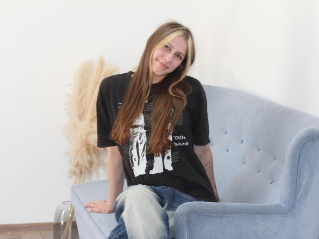 MiaNeit, con cabello largo y castaño, vestida con una camiseta negra y jeans azules, sentada en un sofá azul claro, sonriendo suavemente, con una mano sobre el sofá, y pasto marrón y esponjoso en el fondo..