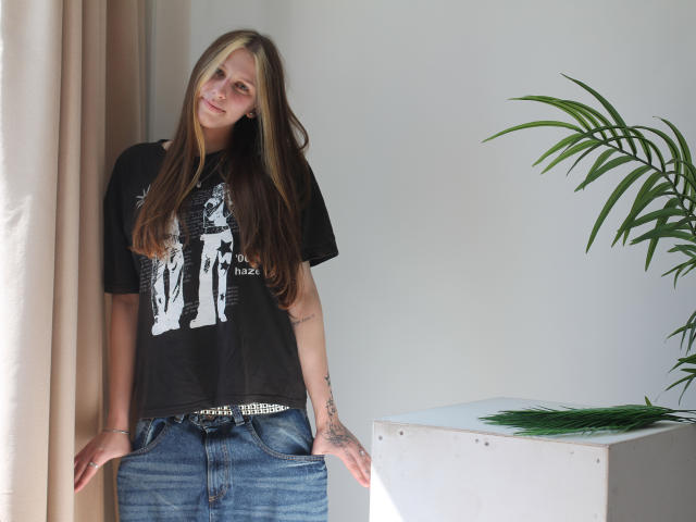 MiaNeit, com cabelos longos loiro-castanhos, camiseta preta e jeans azuis, apoiada na parede, com as mãos nos bolsos, tatuagem no pulso direito, planta em vaso à direita, luz do sol..