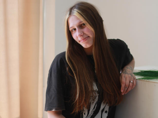 MiaNeit mit langen braunen Haaren, schwarzem T-Shirt, Nasenring und Tattoo auf ihrer rechten Hand, lehnt an einer Wand und lächelt sanft. Ihr Hintern ist nicht sichtbar..