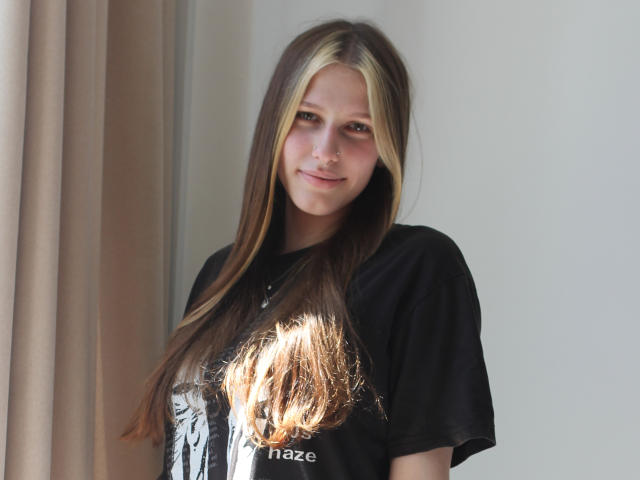 MiaNeit mit langem, braun-blondem Haar, schwarzem T-Shirt, kleinem Nasenring und einem leichten Lächeln, steht in der Nähe von Sonnenlicht. Ihre Brüste sind teilweise im Schatten des Sonnenlichts..