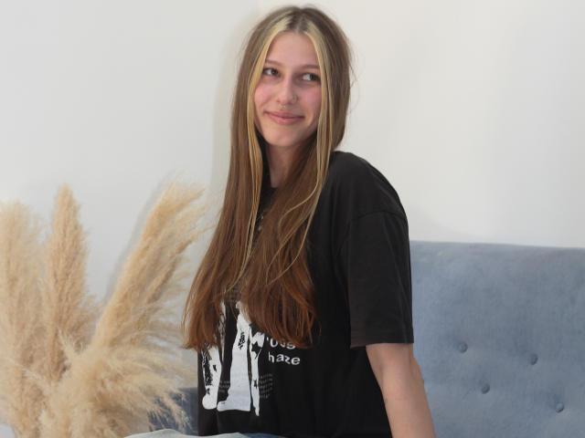 MiaNeit, met lang bruin haar, die licht glimlacht, draagt een zwart T-shirt en staat naast pampa gras, met op de achtergrond een blauw, bekleed hoofdeinde..