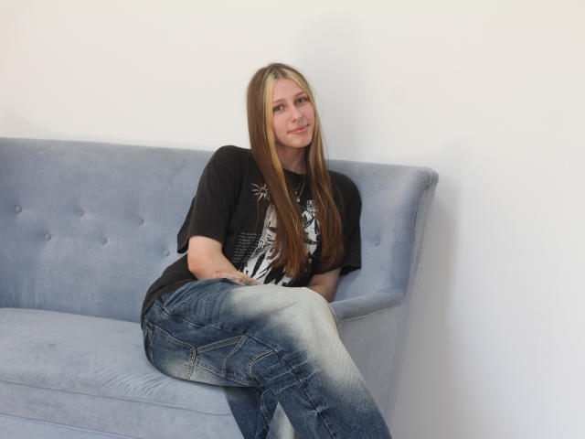 MiaNeit mit langen blonden Haaren, einem schwarzen T-Shirt und blauen Jeans, sitzt auf einem hellblauen, gepolsterten Sofa, lächelt leicht, die Hände auf dem Knie, wirkt lässig und entspannt..