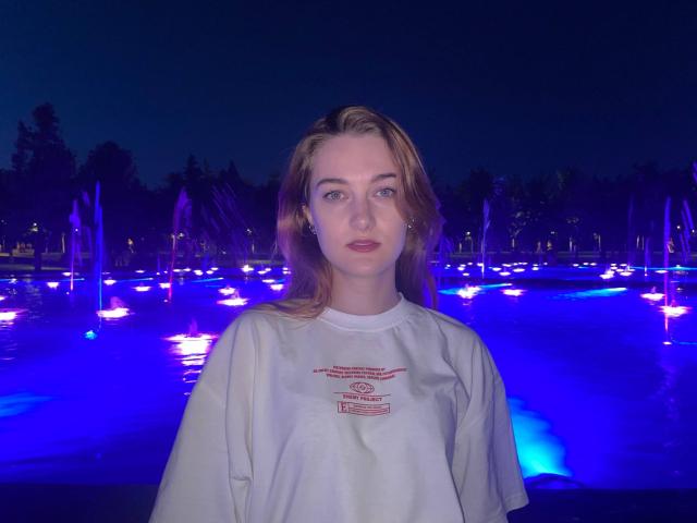 Kirstin Voracek, con la pelle chiara, i capelli castano chiaro e gli occhi azzurri, indossa una camicia bianca e si trova di fronte a un elemento decorativo acquatico viola illuminato, di notte..