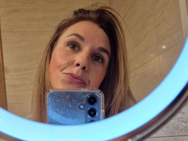 O rosto da PekiPassionX-hot em um selfie no espelho, mostrando olhos castanhos, cabelo loiro liso e lábios rosados, segurando um telefone brilhante, com uma luz de anel azul em primeiro plano..