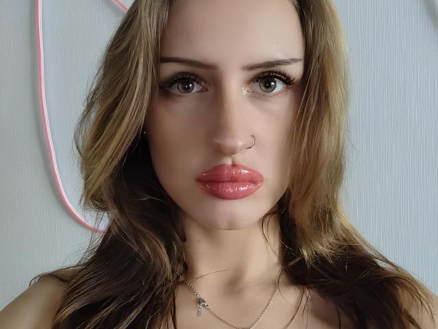 Nicolle Schattner, avec des lèvres roses brillantes, des yeux verts, un anneau au nez et des cheveux bruns ondulés, portant un collier, regardant droit devant la caméra..