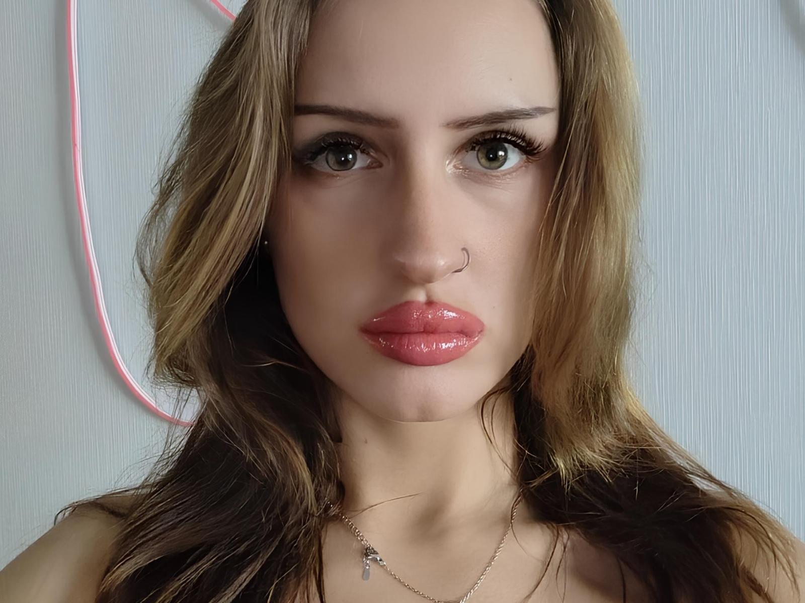 Nicolle Schattner met glanzende, roze lippen, groene ogen, een neusring en bruin, golvend haar, draagt een ketting en kijkt recht in de camera..
