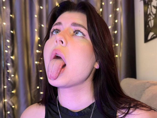 KizzyDepner's face with dark brown hair, blue eyes, and tongue fully out, looking up with mouth open. She's wearing a black top, and has a necklace.