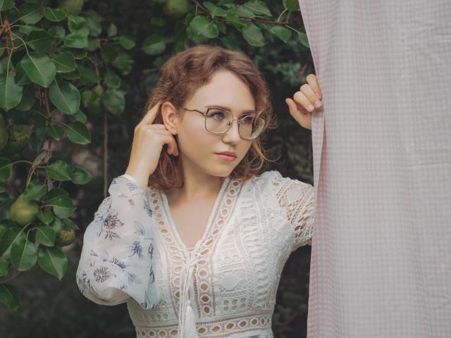 Teresa Birt, con cabello castaño rizado, gafas y un vestido blanco de encaje, sosteniendo una cortina, mirando hacia un lado, de pie cerca de hojas verdes. Los senos están cubiertos por el vestido, la vulva no es visible..