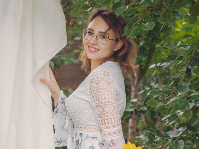 Teresa Birt, avec des lunettes, souriante, tenant un rideau blanc, portant un haut en dentelle, montrant ses seins, dans un jardin, avec des feuilles vertes, des cheveux bruns, et une fleur jaune en bas à droite..
