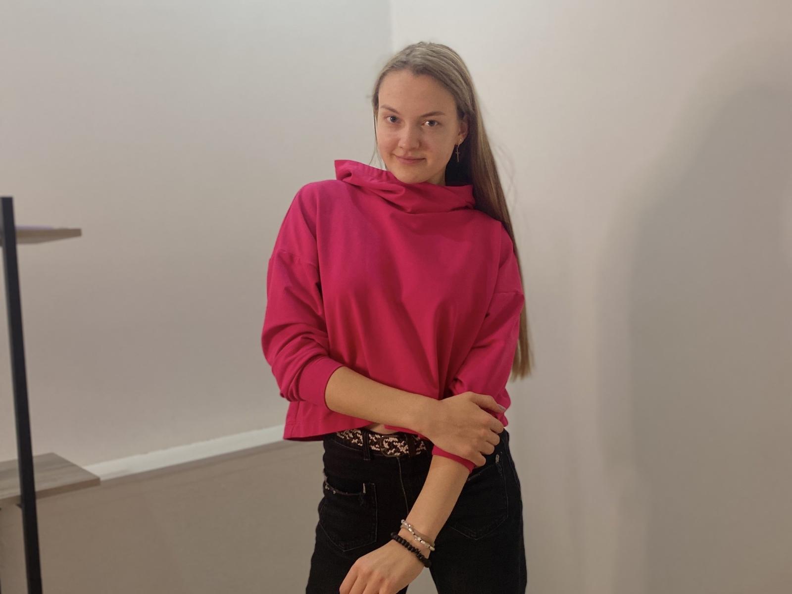 Machelle Gernatt, avec ses longs cheveux blonds, son pull rose à col roulé, son jean noir et sa ceinture léopard, se tient debout, les bras croisés, et sourit légèrement. Elle porte un bracelet à son poignet droit..