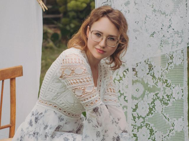 Teresa Birt, met krullend bruin haar, een bril en een witte jurk met kant, leunt op een houten stoel, glimlachend, en laat haar borsten zien door het kantpatroon. Er hangt een kantgordijn op de achtergrond..